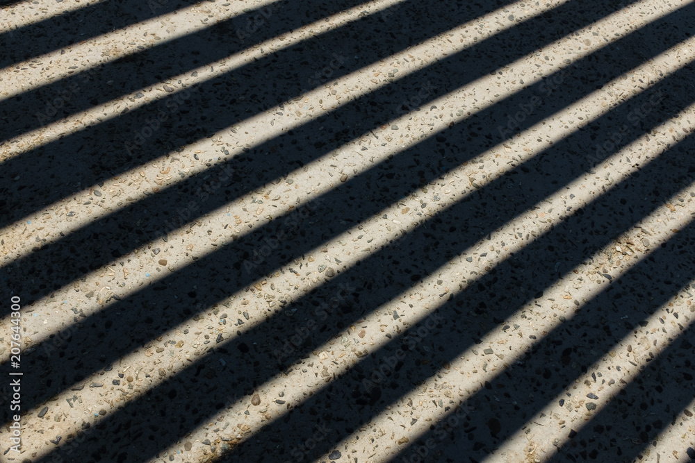 Sunlight shines through wooden fence and makes geometric stripes shadows on the ground. Shadows of wooden fence on the ground. Fence shadows texture