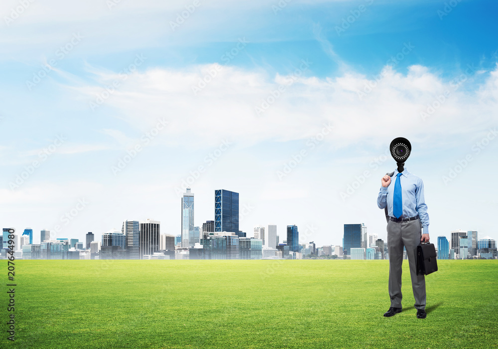 Camera headed man standing on green grass against modern cityscape Camera headed man standing on green grass against modern cityscape