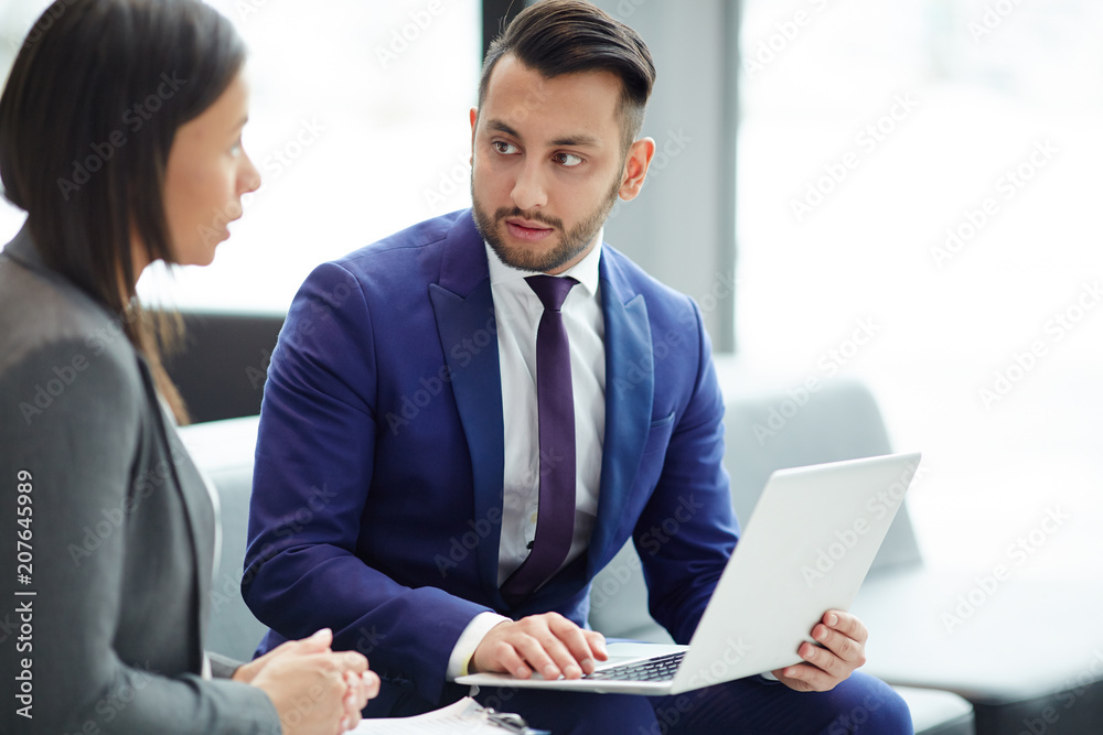 Young businessman with laptop looking at colleague while making presentation or discussing online data Young businessman with laptop looking at colleague while making presentation or discussing online data