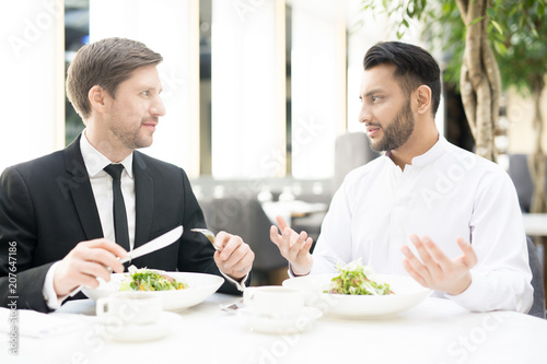 Two young intercultural businessmen sitting by served table during business lunch and communicating