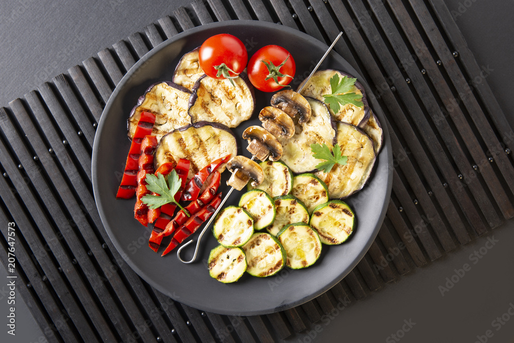 Grilled vegetables: eggplant, zucchini, pepper. Skewer with mushrooms. On a black plate and a black background. Toned.