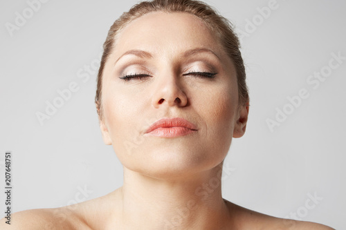 Handsome pretty woman with clean skin, natural make-up, and closed eyes holding hands on her face on gray background. Close up portrait. Medical and cosmetic facial skin care concept
