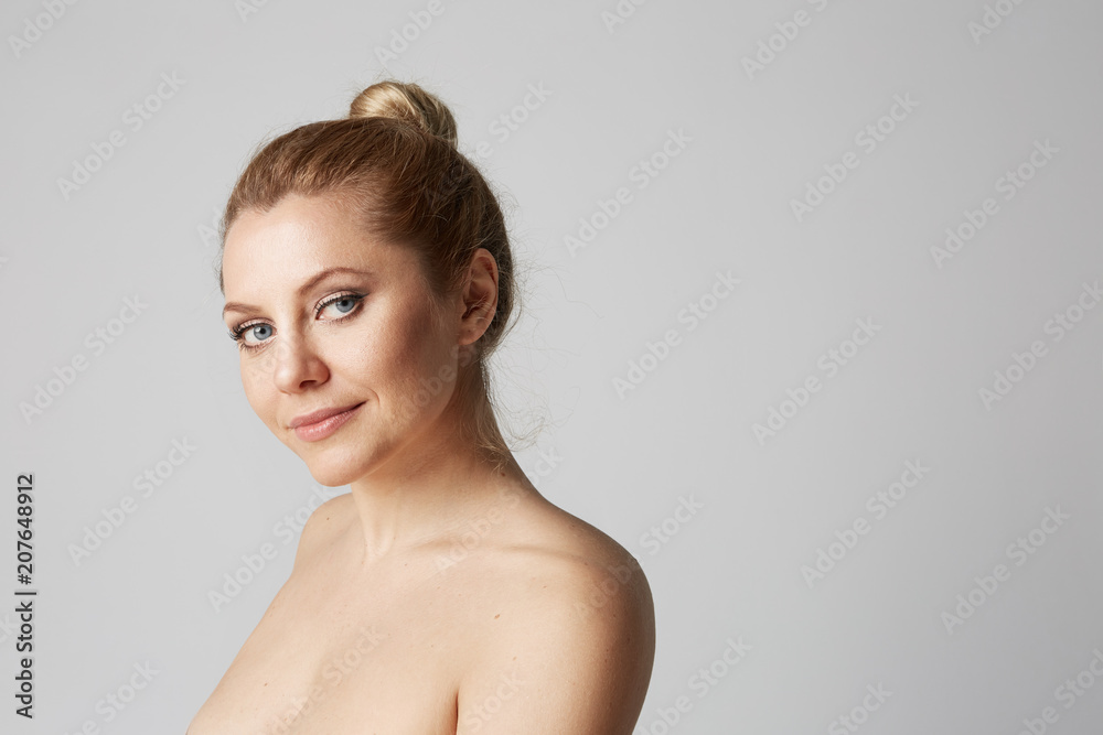Young beautiful girl with blonde hair fixed behind, big eyes, thick eyebrows and naked shoulders smiling on gray background.Medical and cosmetic facial skin healthcare concept.Copy paste space