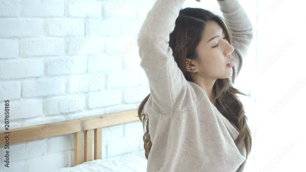 Happy beautiful young Asian woman waking up in morning, sitting on bed, stretching in cozy bedroom, looking through window. Funny asian woman after wake up. Asia woman is stretching and smiling.