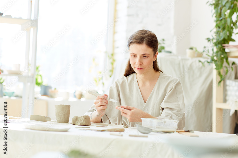 Young female in workwear sitting by table in her workshop and making earthenware Young female in workwear sitting by table in her workshop and making earthenware