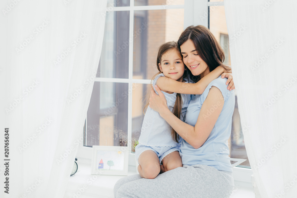 Lovely small child with pleasant appearance embraces her mother, expresses love and good feeling or attitude, sit on window sill, enjoy domestic atmosphere and togetherness. Parenthood concept Lovely small child with pleasant appearance embraces her mother, expresses love and good feeling or attitude, sit on window sill, enjoy domestic atmosphere and togetherness. Parenthood concept