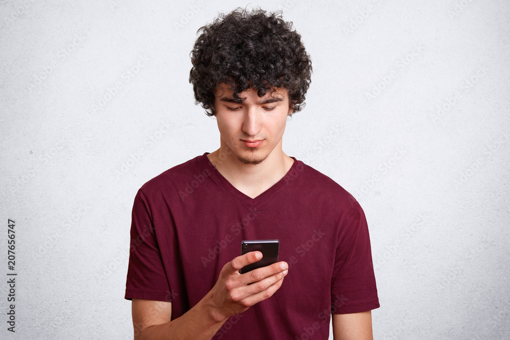 Good looking guy with concetrated look in mobile phone, recieves message with offer to go out, wears casual t shirt, poses against white concrete wall. Curly man uses modern technologies indoor Good looking guy with concetrated look in mobile phone, recieves message with offer to go out, wears casual t shirt, poses against white concrete wall. Curly man uses modern technologies indoor