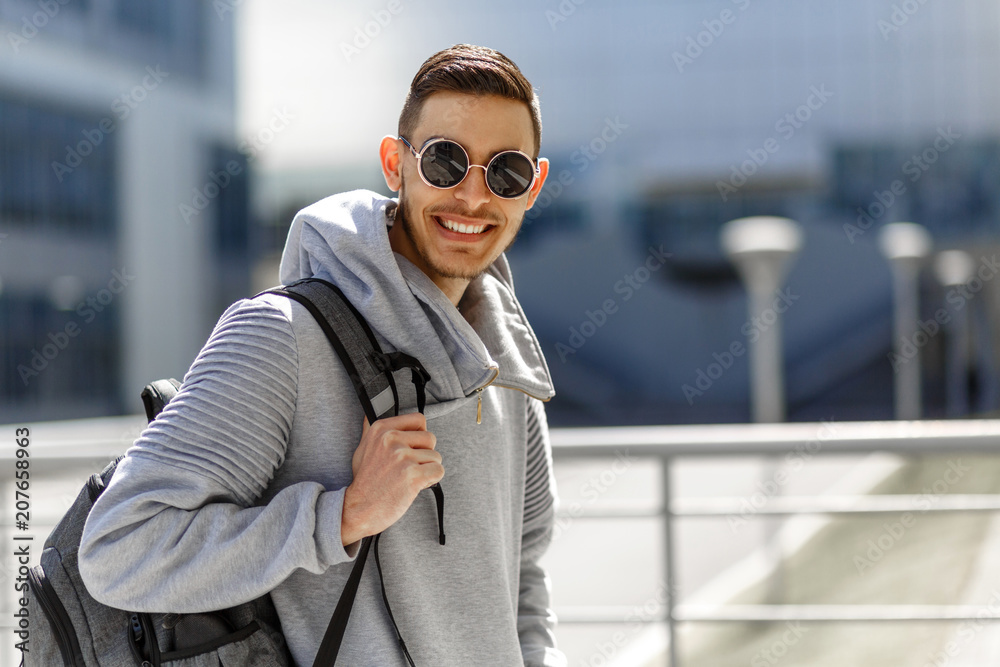 Handsome stylish Caucasian man wearing trendy sunglasses posing against street background with backpack on his shoulders. Traveler man view from above in the city. Lifestyle fashion urban concept.