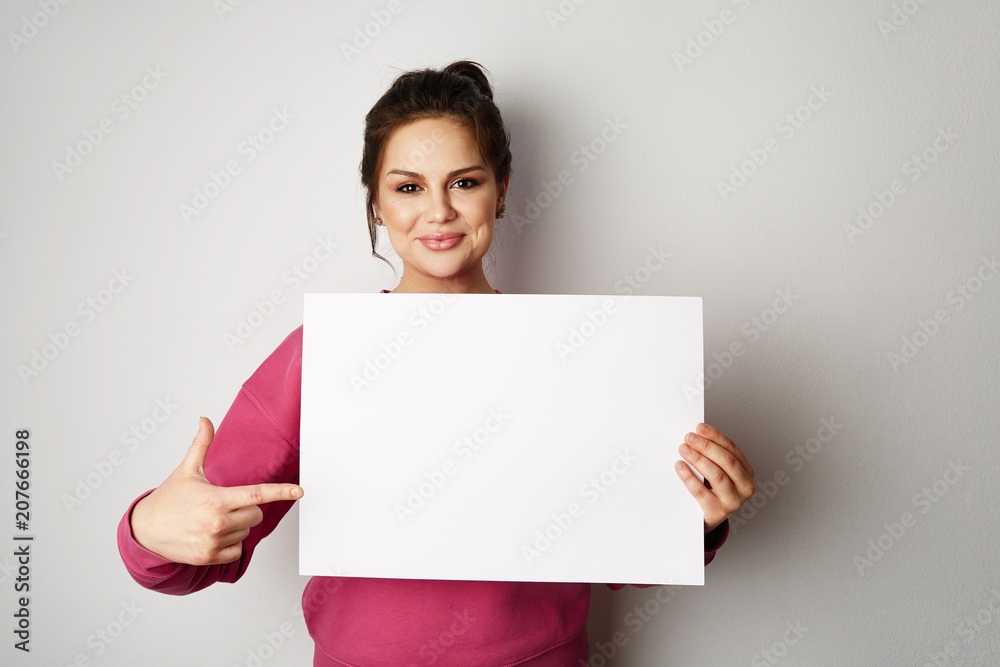 Pretty smiling woman in pink hoody holding empty blank board isolated on the gray background - obrazy, fototapety, plakaty 
