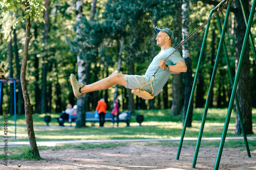 Adult stylish man in glasses riding on swing in city park on playground for children in summer. Happy guy remember childhood. Male person on attraction enjoying up and down motion. Odd boy outdoor. Adult stylish man in glasses riding on swing in city park on playground for children in summer. Happy guy remember childhood. Male person on attraction enjoying up and down motion. Odd boy outdoor.