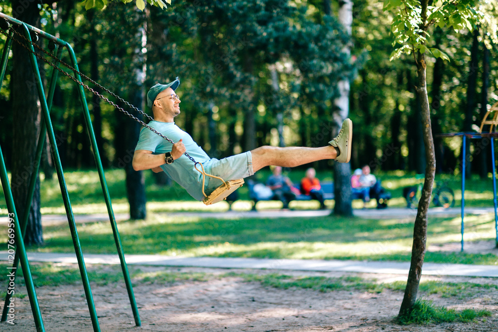 Adult stylish man in glasses riding on swing in city park on playground for children in summer.  Happy guy remember childhood.  Male person on attraction enjoying up and down motion.  Odd boy outdoor.