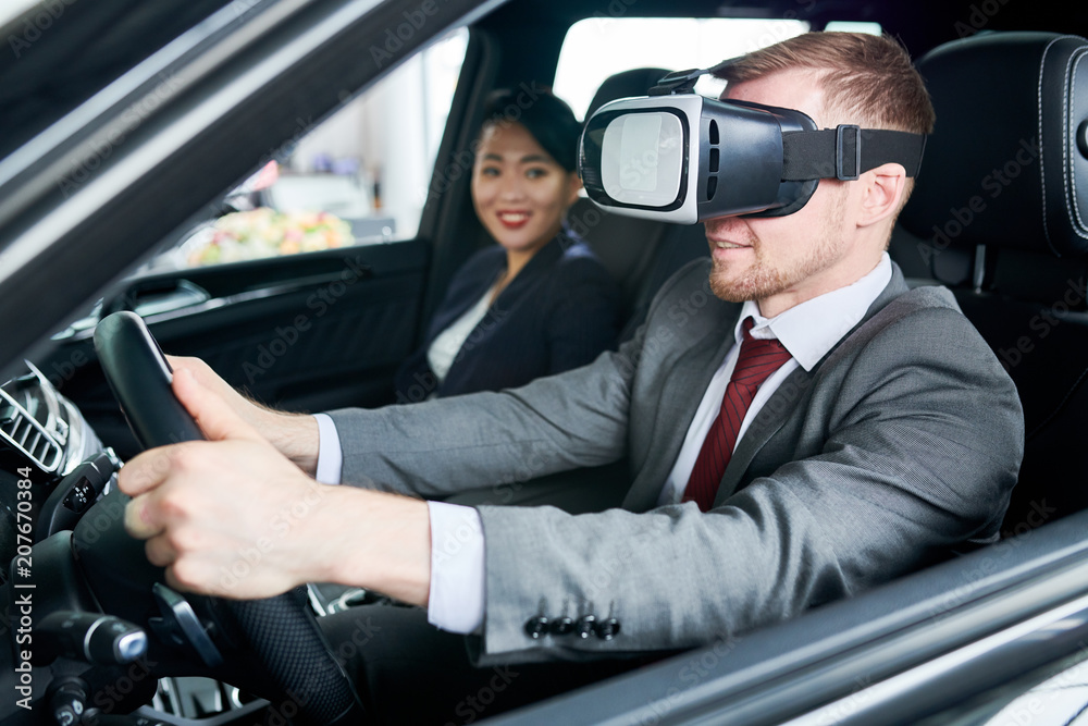 Bearded man wearing classical suit using VR headset while learning to drive car, pretty Asian instructor assisting him