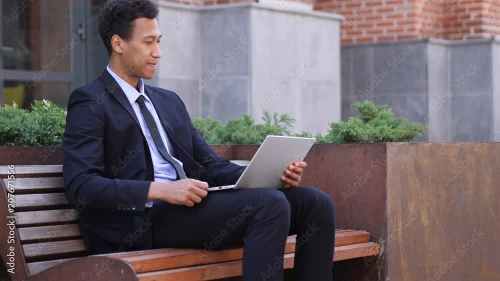 Excited African Businessman Celebrating Success on Laptop Sitting on bench Excited African Businessman Celebrating Success on Laptop Sitting on bench