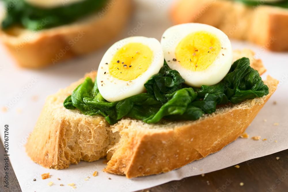 Crostini roasted bread slices with cooked spinach leaves and hard boiled quail eggs seasoned with black pepper, photographed with natural light (Selective Focus, Focus on the front of the egg yolks)