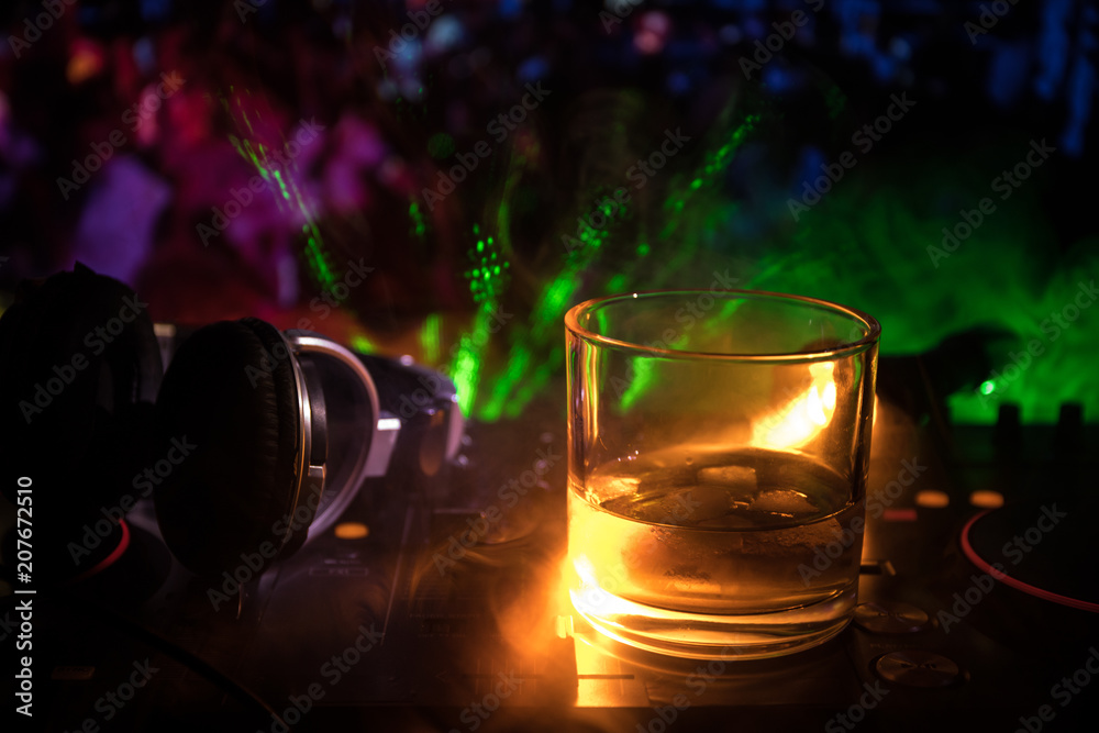 Glass with whisky with ice cube inside on dj controller at nightclub. Dj Console with club drink at music party in nightclub with disco lights. Glass with whisky with ice cube inside on dj controller at nightclub. Dj Console with club drink at music party in nightclub with disco lights.