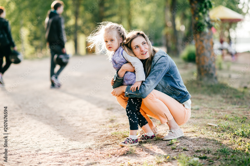 Fototapeta premium Happy smiling mother hugging her lovely little daughter outdoor. Lifestyle family. Adult cheerful female parent playing with her beautiful emotional child at nature in summer. Positive people faces.