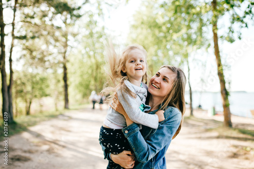Happy smiling mother hugging her lovely little daughter outdoor. Lifestyle family. Adult cheerful female parent playing with her beautiful emotional child at nature in summer. Positive people faces.