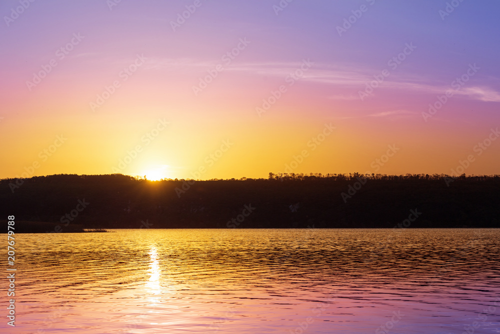 Dramatic sunset over river with orange and purple colors.Sun goes down over pond and forest silhouette. Beautiful nature background Dramatic sunset over river with orange and purple colors.Sun goes down over pond and forest silhouette. Beautiful nature background