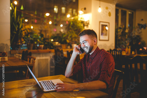 Young handsome Caucasian man with beard and toothy smile in red shirt works behind laptop, hands on keyboard sitting at wooden table. Uses calls on mobile phone. In evening at the coffee shop.