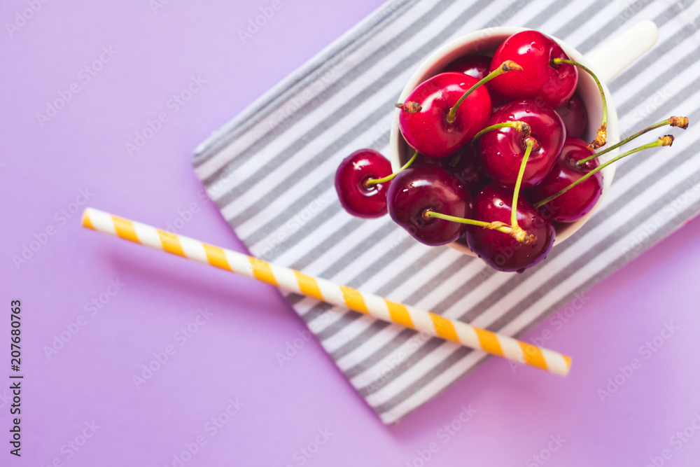 Φωτογραφία Close up and top view of fresh red cherries in a white cup on a gray and white stripped cloth, a yellow and white straw on pink or purple background