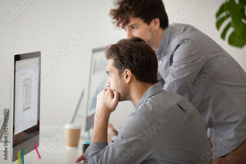 Caucasian young male mentor helping colleague with computer work during company meeting, consulting on software issues, explaining and coaching. Concept of collaboration, cooperation