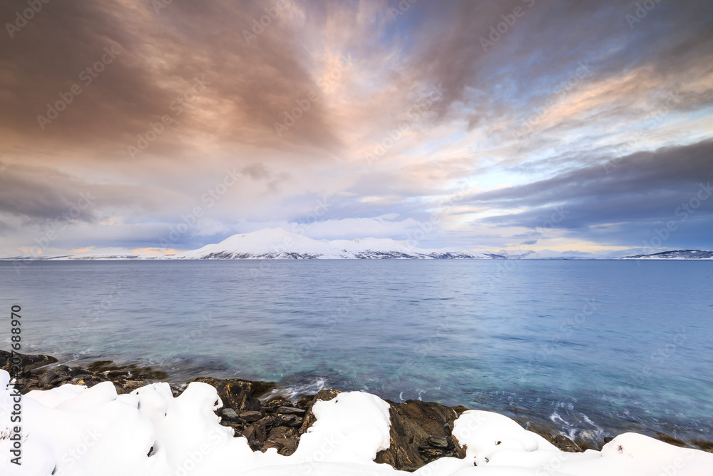 Sunset at the lakeside with rocks of a fjord during low tide in a snowy winter landscape.