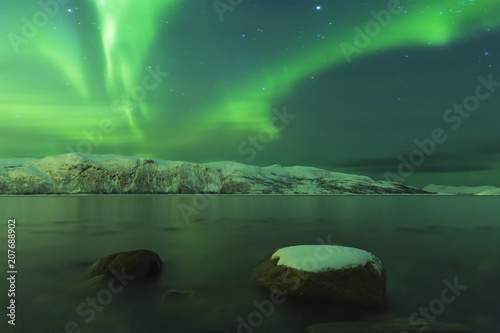 Northern lights Aurora borealis at the lakeside with rocks of a fjord during low tide in a snowy winter landscape with mountains