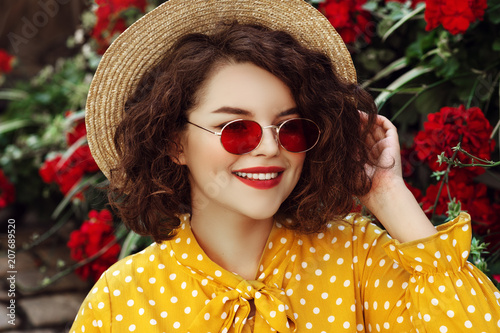 Outdoor close up portrait of young beautiful stylish happy smiling curly girl wearing red narrow oval sunglasses, hat, yellow dress, posing in street with flowers. Summer fashion concept
