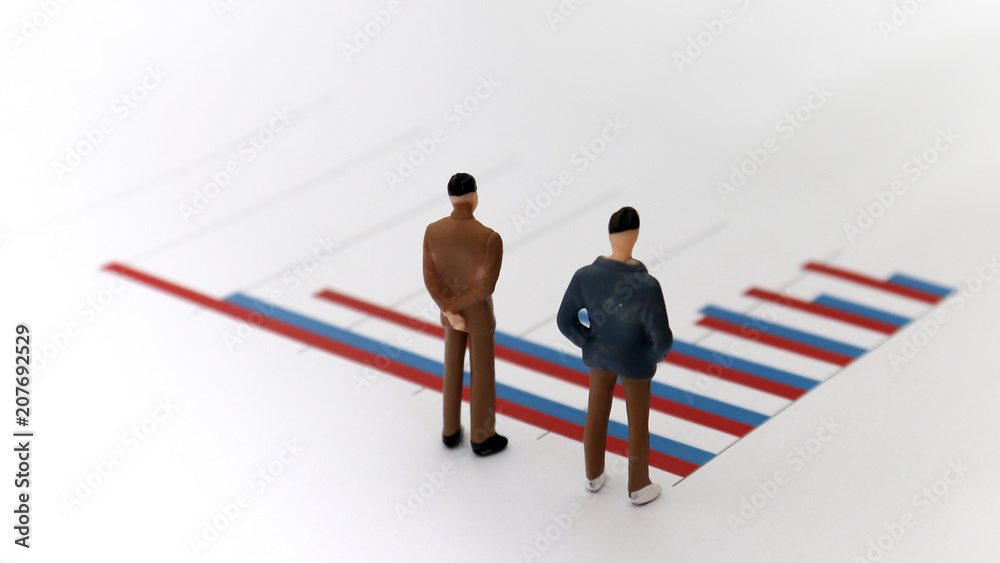 Fototapeta premium Appearance from behind of two miniature men standing on a bar graph.