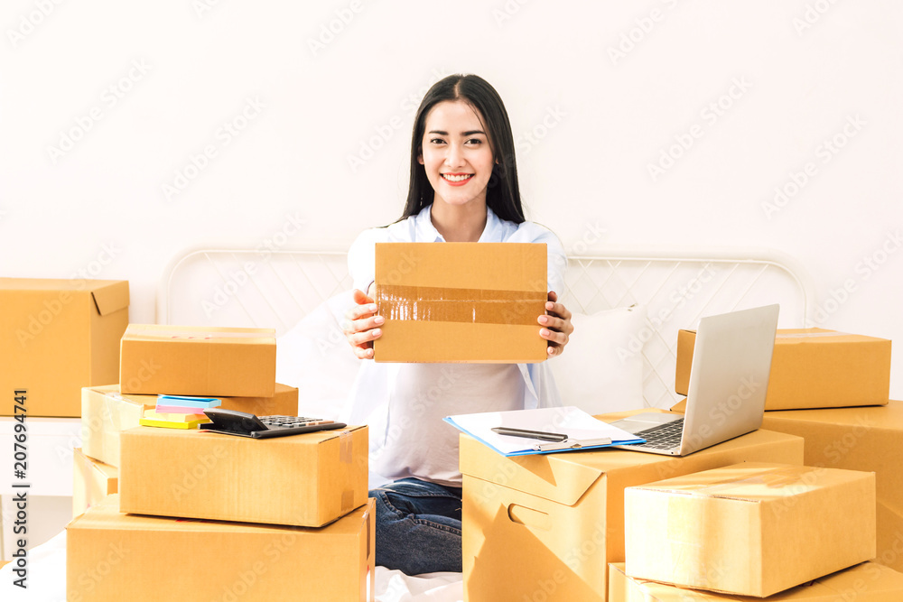 Young woman freelancer working and holding cardboard box on bed at home - SME business online and delivery concept Young woman freelancer working and holding cardboard box on bed at home - SME business online and delivery concept