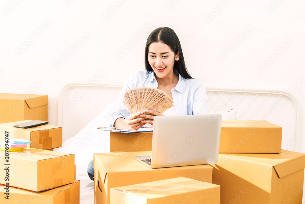 Young woman freelancer working and holding money with cardboard box on bed at home - SME business online and delivery concept Young woman freelancer working and holding money with cardboard box on bed at home - SME business online and delivery concept