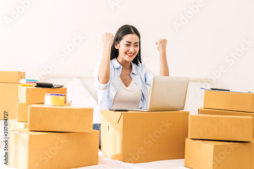 Successful young woman freelancer with arms up working and use laptop computer with cardboard box on bed at home - SME business online and delivery concept