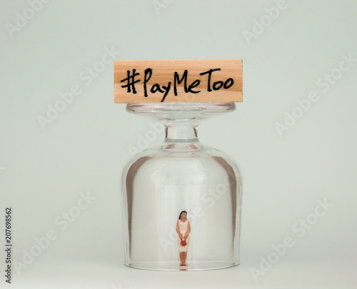 #PayMeToo as a new social movement. A miniature woman in a glass cup and a wooden block with #PayMeToo written on top of a glass cup. photo