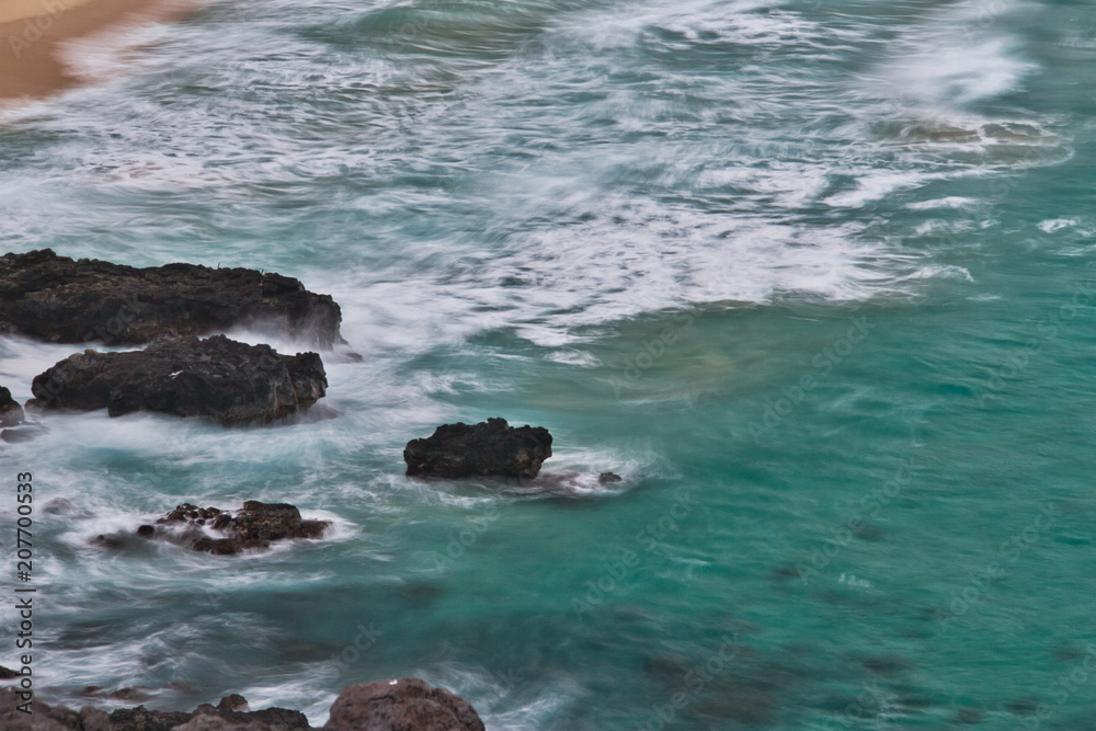 Long exposure image looking down on waves crashing against lava rocks on a beach.