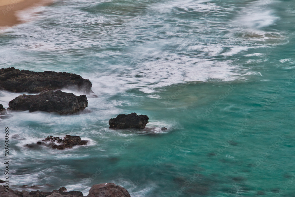 Long exposure image looking down on waves crashing against lava rocks on a beach. Long exposure image looking down on waves crashing against lava rocks on a beach.