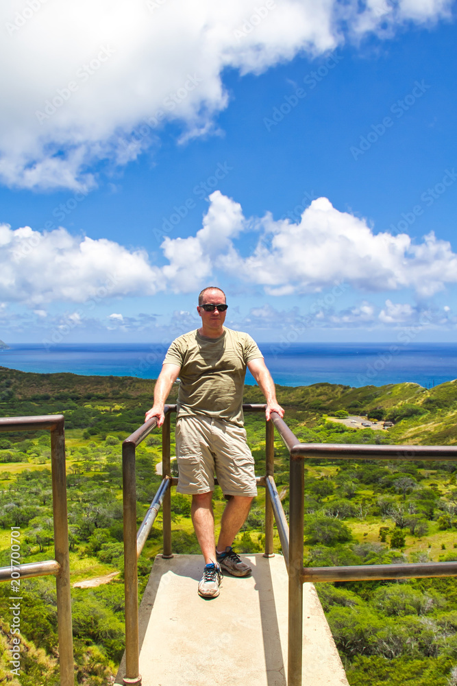 A male hiker resting at an overlook in Hawaii and enjoying the lush green vegetation and the blue ocean and sky.
