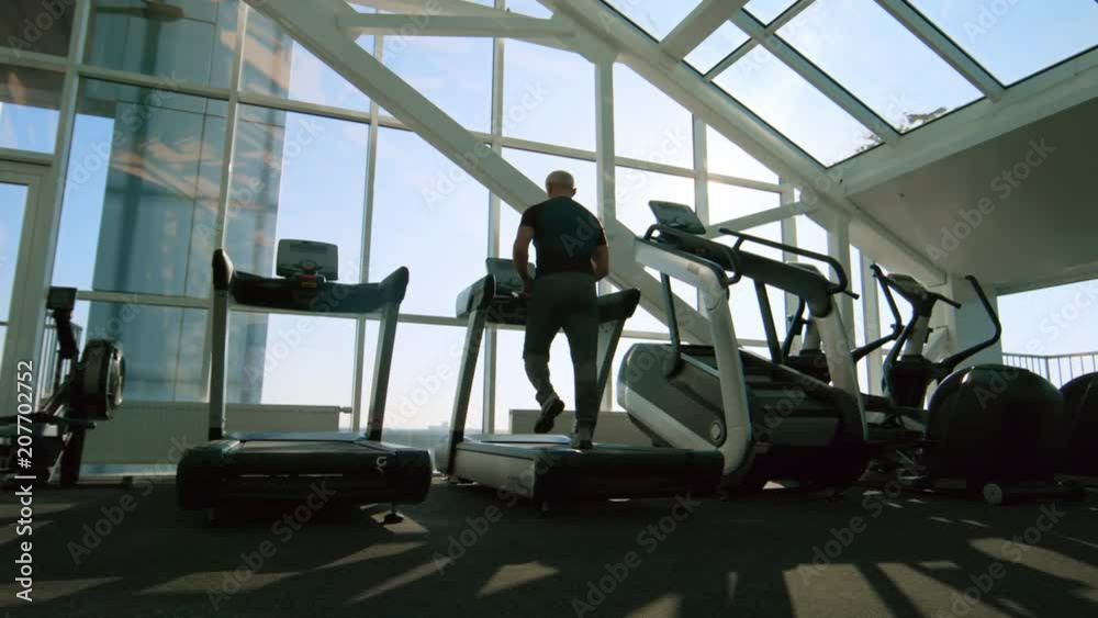 PAN with rear view of senior man with grey hair walking on treadmill during workout in gym with panoramic windows PAN with rear view of senior man with grey hair walking on treadmill during workout in gym with panoramic windows