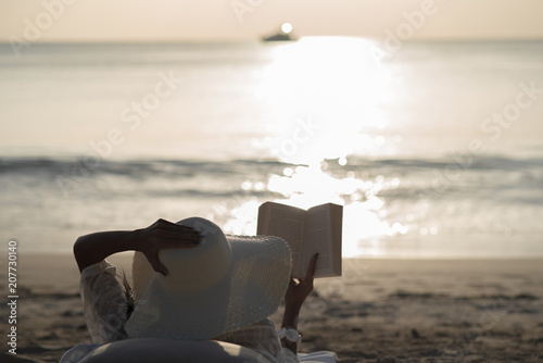 Fototapeta Naklejka Na Ścianę i Meble -  Woman with white hat sit on the beach reading the booking during sunset.