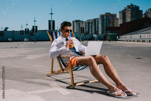 handsome young businessman in shorts working with laptop while sitting on sun lounger on parking and drinking cocktail