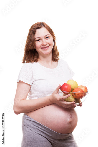 Pregnant woman smiling to the camera holds an glass bowl with fruits in her hands. Isolated over white background