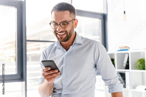 Image closeup of smiling employer guy in white shirt standing in office room near big window, and using smartphone for work photo