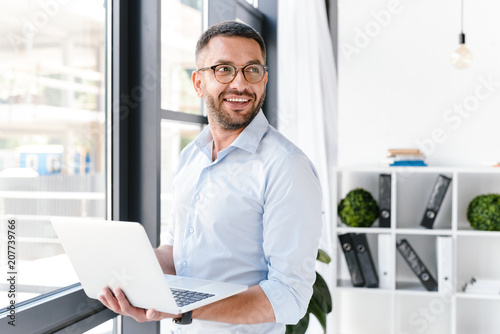 Image of office worker man wearing white shirt looking aside, while holding silver laptop during work in business centre