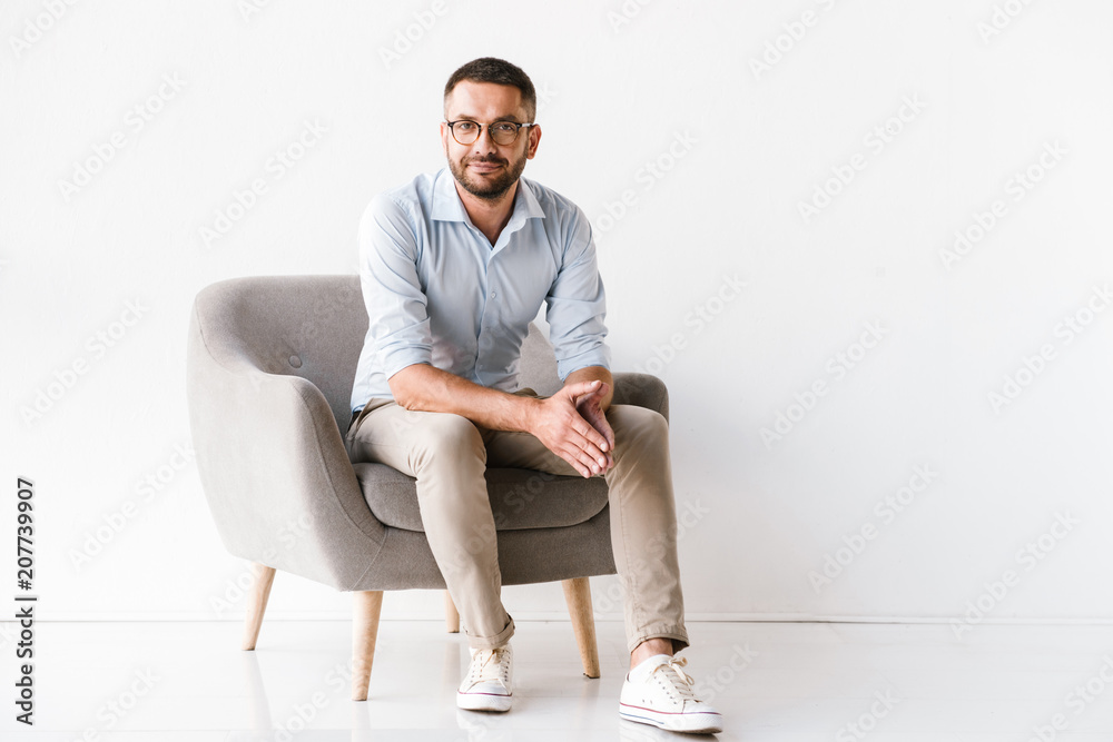 Horizontal portrait of attractive caucasian guy wearing stylish formal clothing sitting in armchair and looking at camera, isolated over white background - obrazy, fototapety, plakaty 