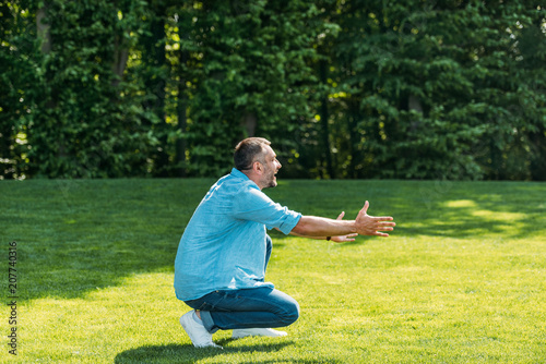 side view of happy man with open arms crouching and looking away in park