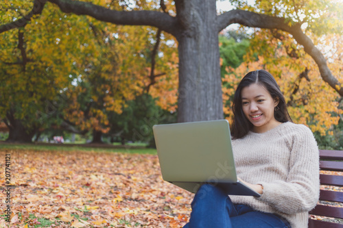 Asian beautiful smiling woman working with laptop while sitting in garden with green grass and falling leaf in autumn.Concept of people using technology.