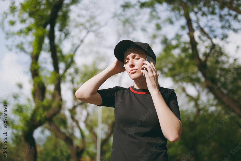 Young athletic tired beautiful brunette girl in black uniform and cap talking on mobile phone during training, keeping hand near head in city park outdoors. Fitness, healthy lifestyle concept. Young athletic tired beautiful brunette girl in black uniform and cap talking on mobile phone during training, keeping hand near head in city park outdoors. Fitness, healthy lifestyle concept.