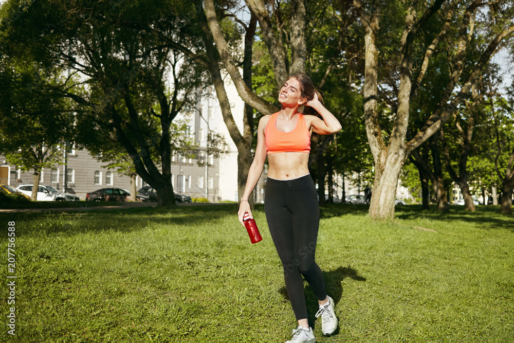 Full lenght portrait of slim athletic young woman with pony tail walking along grass in park, holding bottle of water, feeling happy after successful jogging workout, enjoying hot sunny summer day
