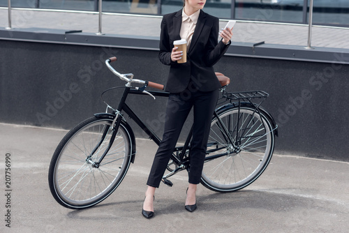 partial view of businesswoman with coffee to go and smartphone standing near retro bicycle on street