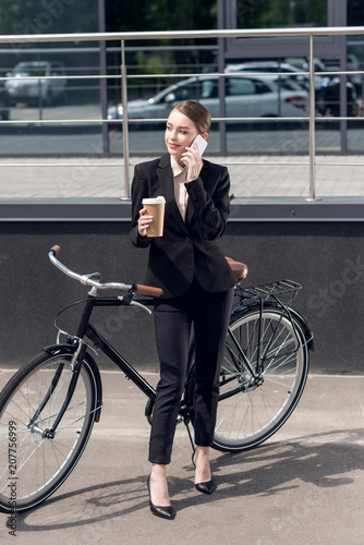 young businesswoman with coffee to go talking on smartphone while standing near retro bicycle on street