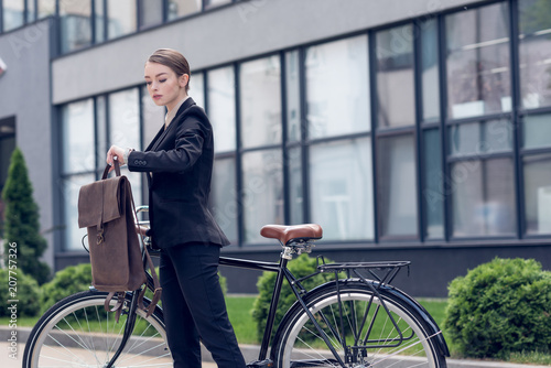 Fototapeta Naklejka Na Ścianę i Meble -  portrait of young businesswoman with briefcase and retro bicycle checking time on street